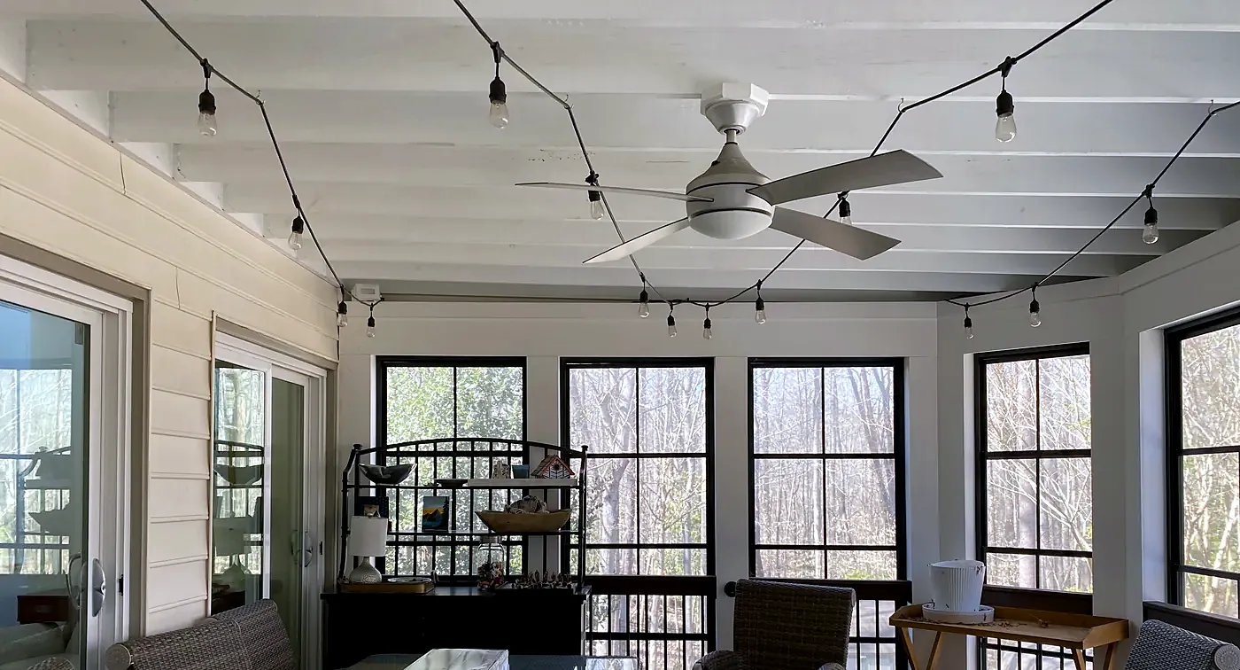 White low-profile ceiling fan on a sunroom ceiling with string lights, black window frames and wicker chairs