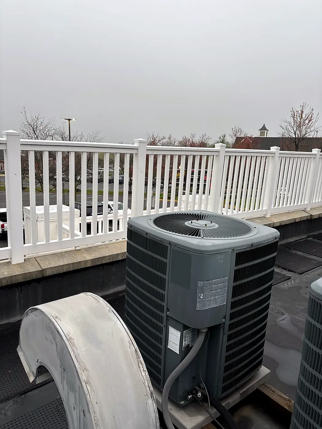 Air conditioner condenser on a flat rooftop deck next to a white vinyl railing under an overcast sky