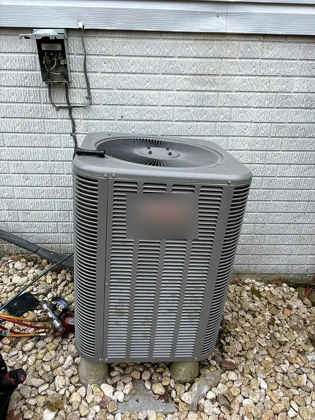 Condenser on blocks over a river-rock bed against a light gray block wall with a disconnect box above
