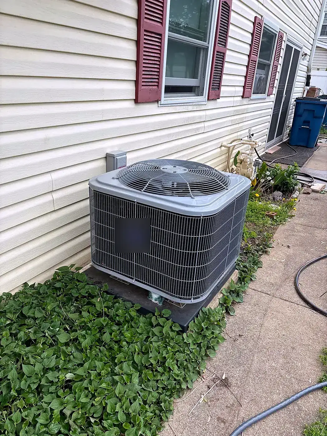 New AC condenser beside a cream vinyl-sided house with maroon shutters, groundcover around the pad