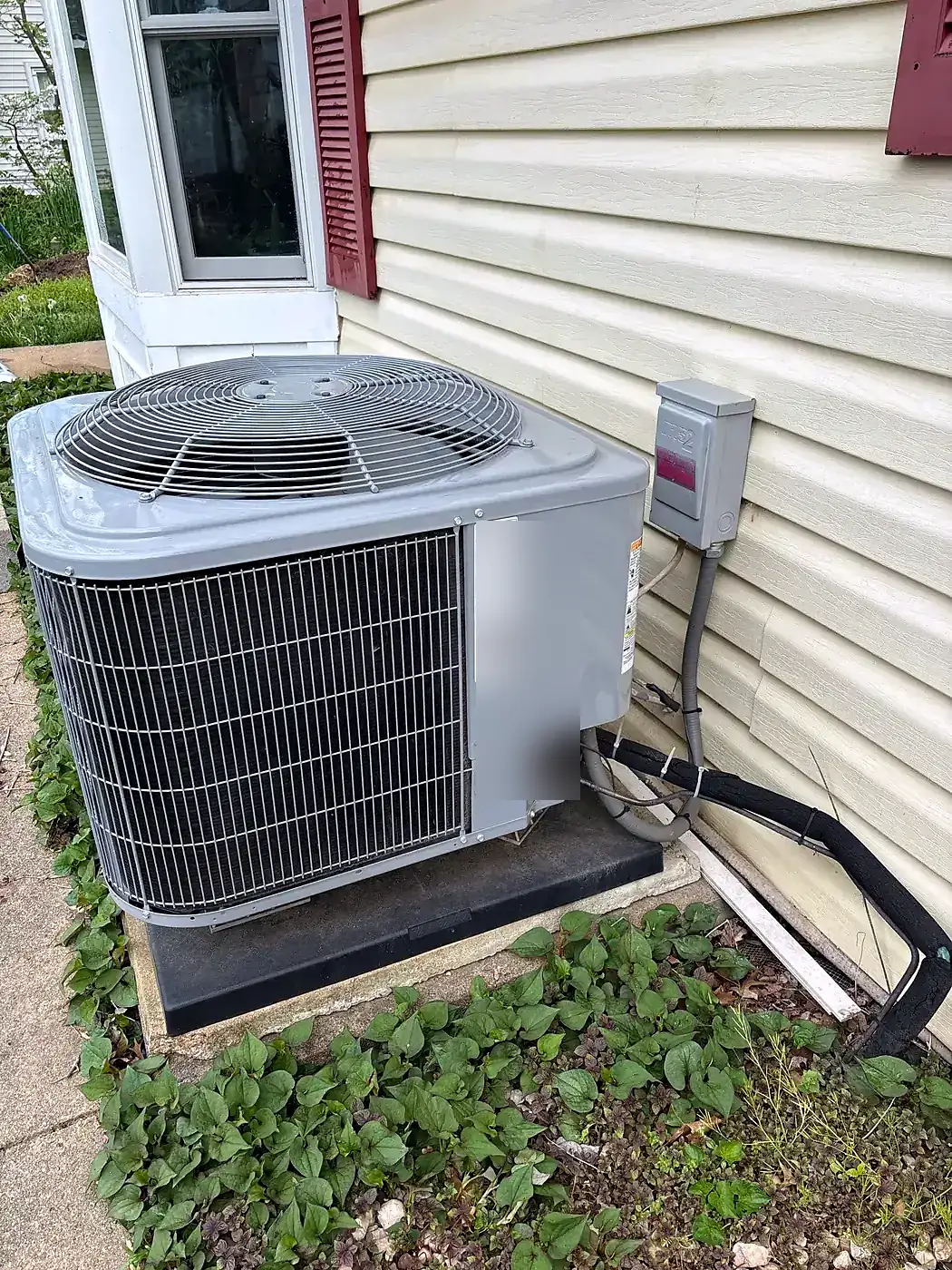 AC condenser on a black pad under a window with a gray disconnect box mounted on cream siding