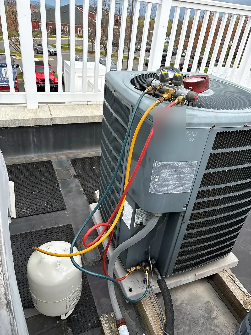 Gray air conditioner condenser on a rooftop deck with gauge hoses attached and a refrigerant tank beside it