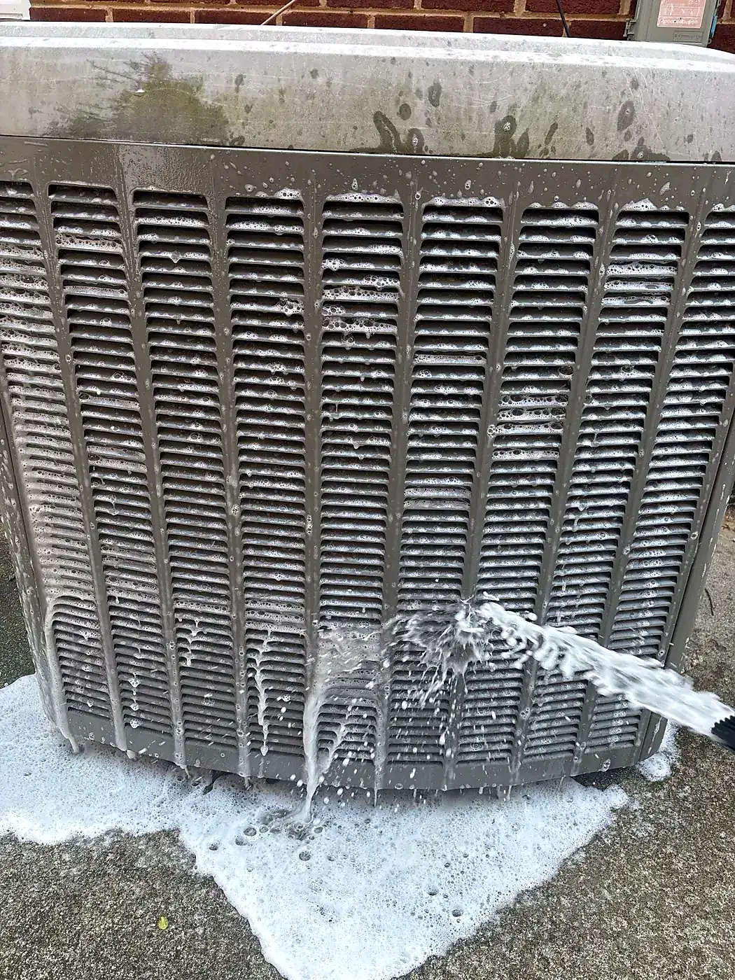 Close-up of outdoor condenser coil fins covered in white foaming cleaner, foam pooling on the pavement below