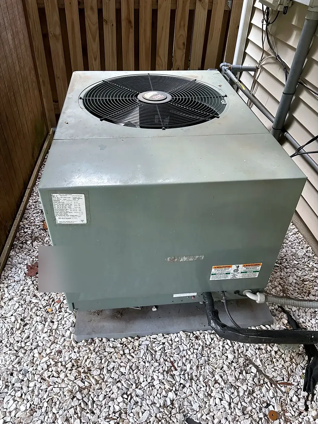 Older green air conditioner condenser on white gravel next to a house wall and a wood fence