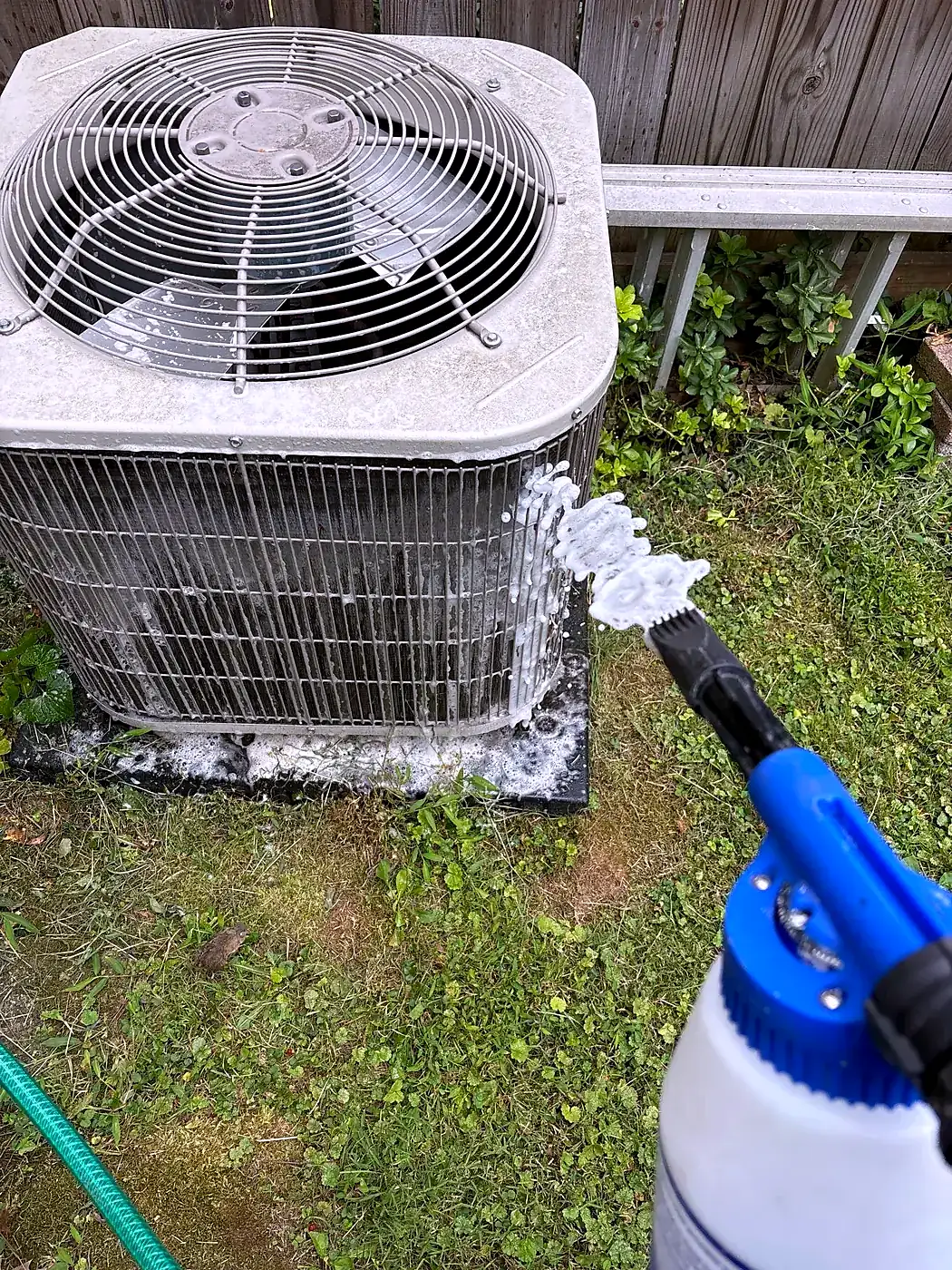 Pump sprayer applying cleaner to an outdoor AC condenser coil in a grassy yard with a garden hose nearby