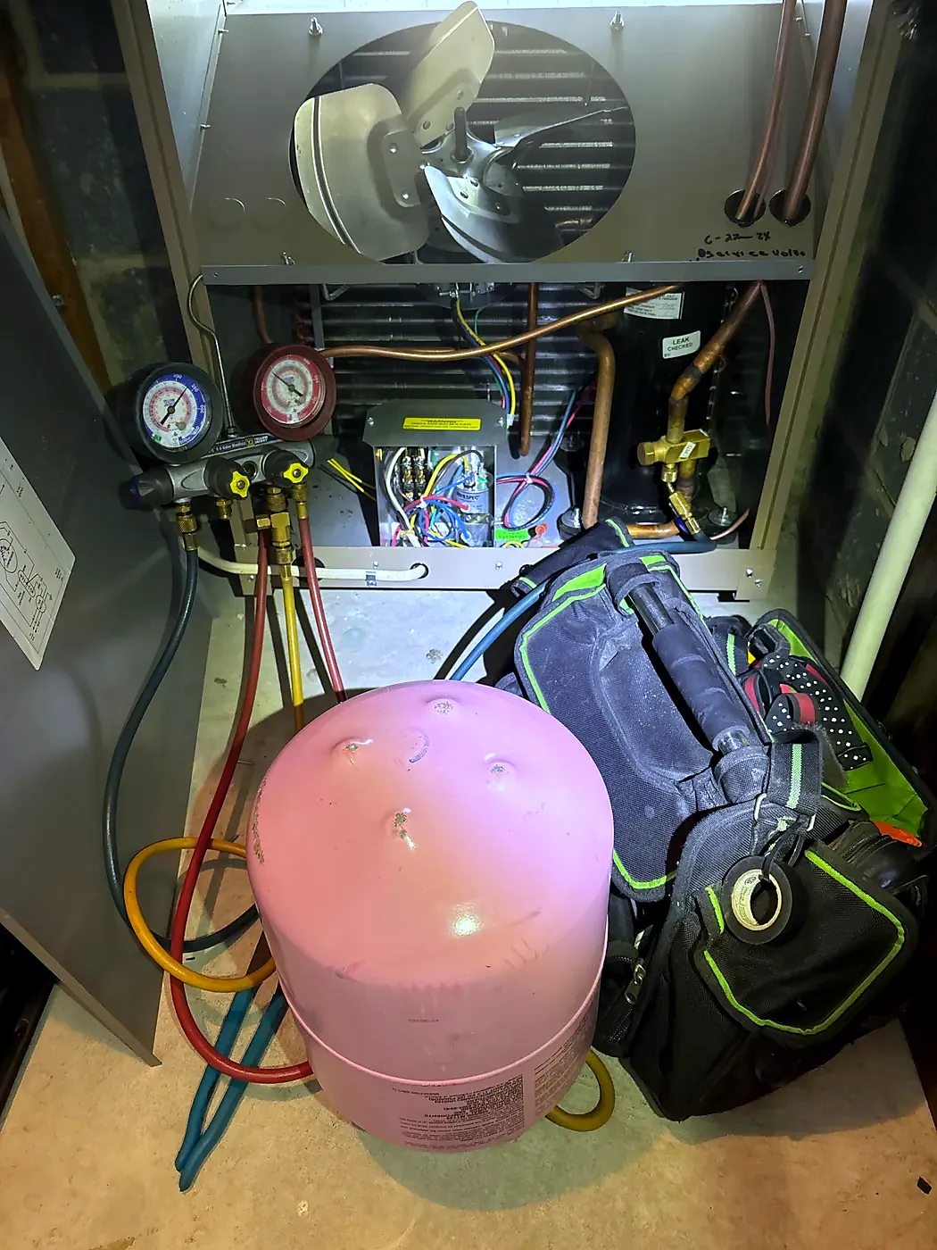Open furnace cabinet with manifold gauges, a pink refrigerant cylinder and a tool bag on the floor in front