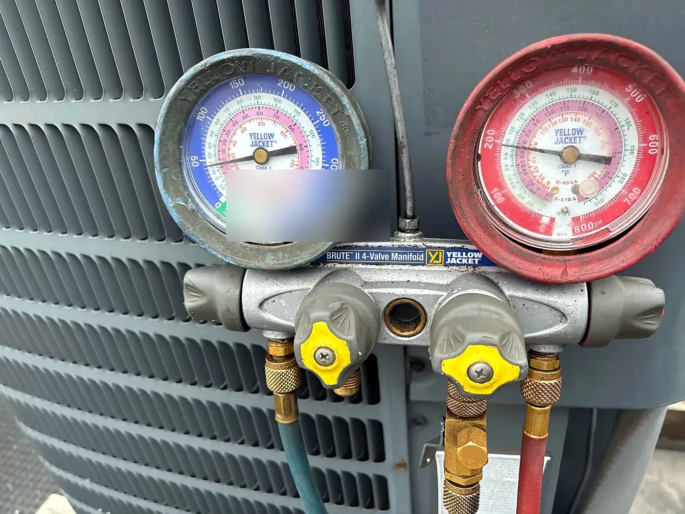 Close-up of a red and blue manifold gauge set hanging on the gray louvered side of an AC condenser