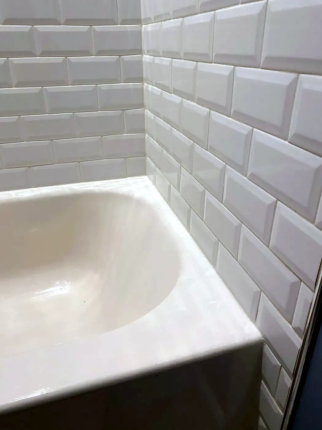 Corner of a white bathtub with white beveled subway tile and a fresh white caulk seam