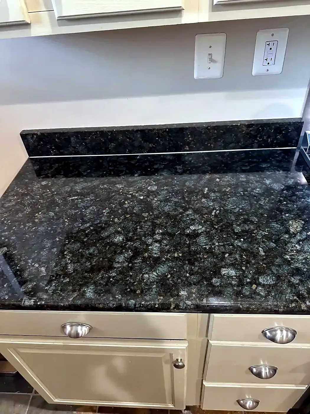 Speckled dark granite counter and backsplash above cream drawers, new caulk line at the joint