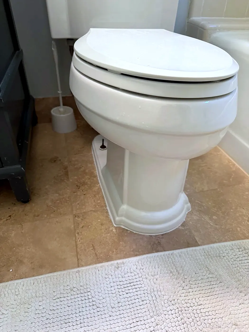 White toilet base sealed with caulk to a tan travertine tile floor, bath rug in front