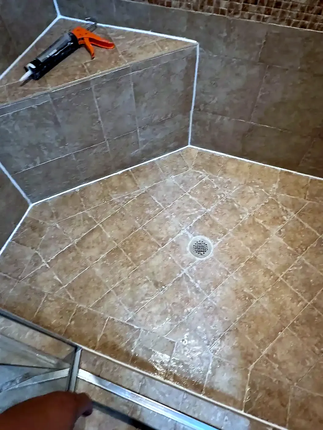 Tan tile shower floor in a diamond pattern with fresh grout, center drain and a tiled bench with a caulk gun on it