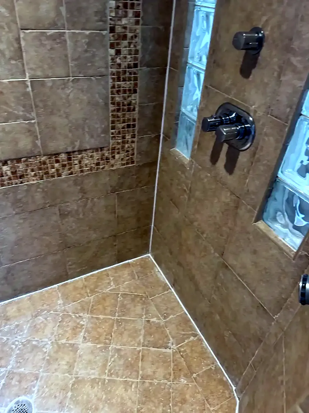Tan tiled shower stall with a mosaic accent band, glass block window and chrome shower valve, freshly regrouted
