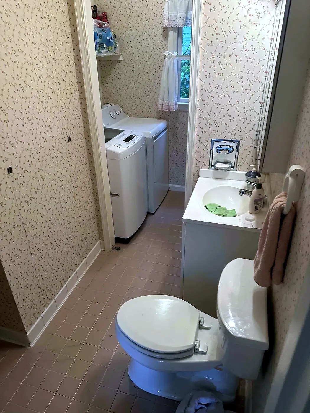 Narrow older bathroom with floral wallpaper, small vanity, toilet, and washer and dryer at the end