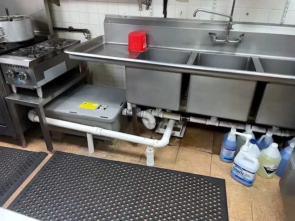 Three-compartment stainless steel sink with white PVC drain piping in a commercial kitchen, rubber floor mat below