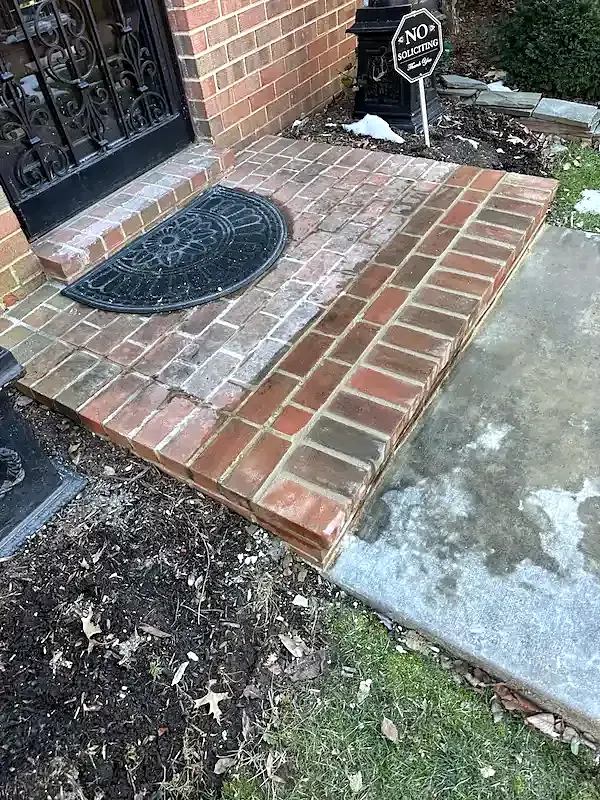Angled view of a red brick front step meeting a gray concrete walkway, doormat at the door