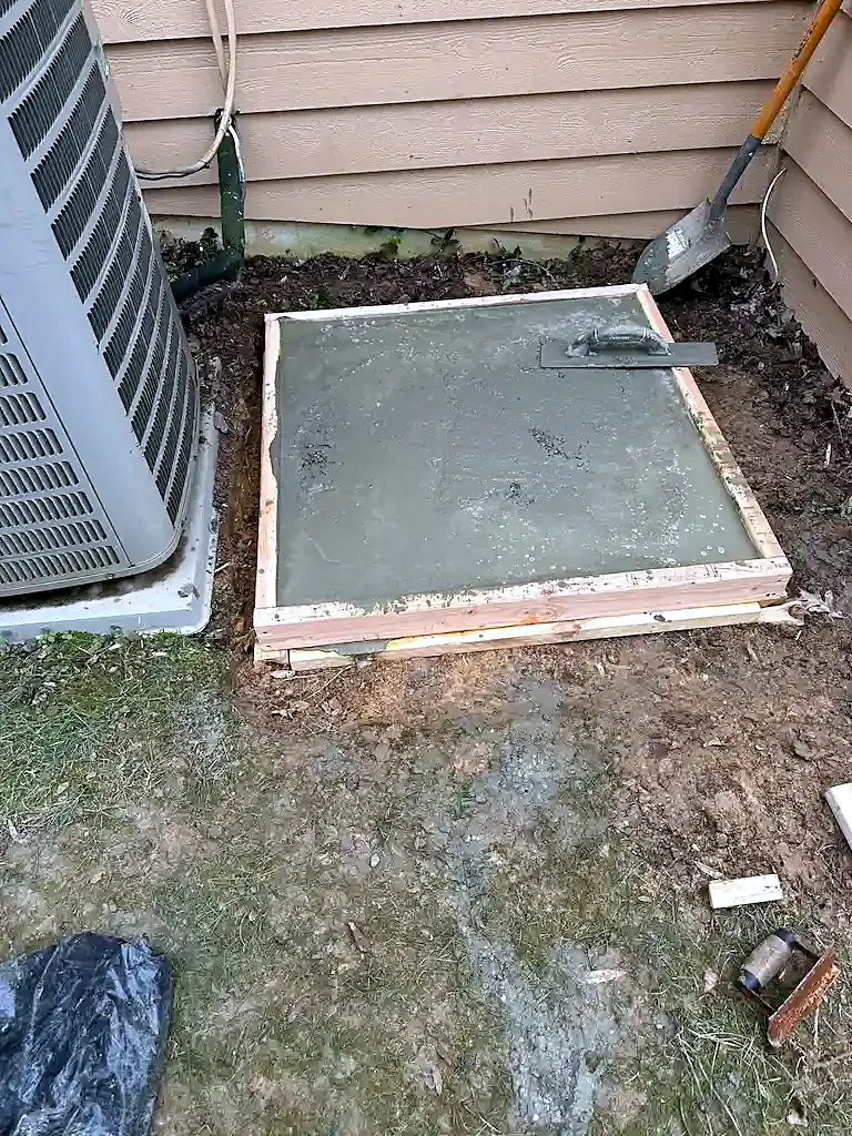 Freshly poured concrete pad in wood forms next to an AC condenser, trowel resting on the surface