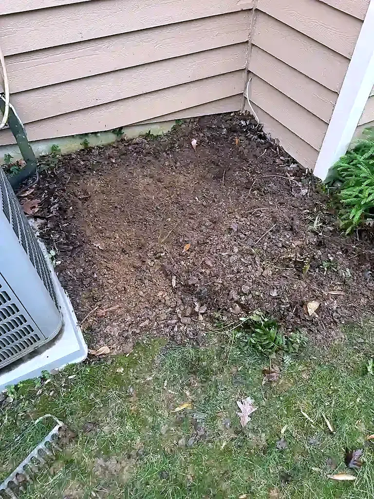Bare dirt patch beside an AC condenser at the corner of a house with tan siding