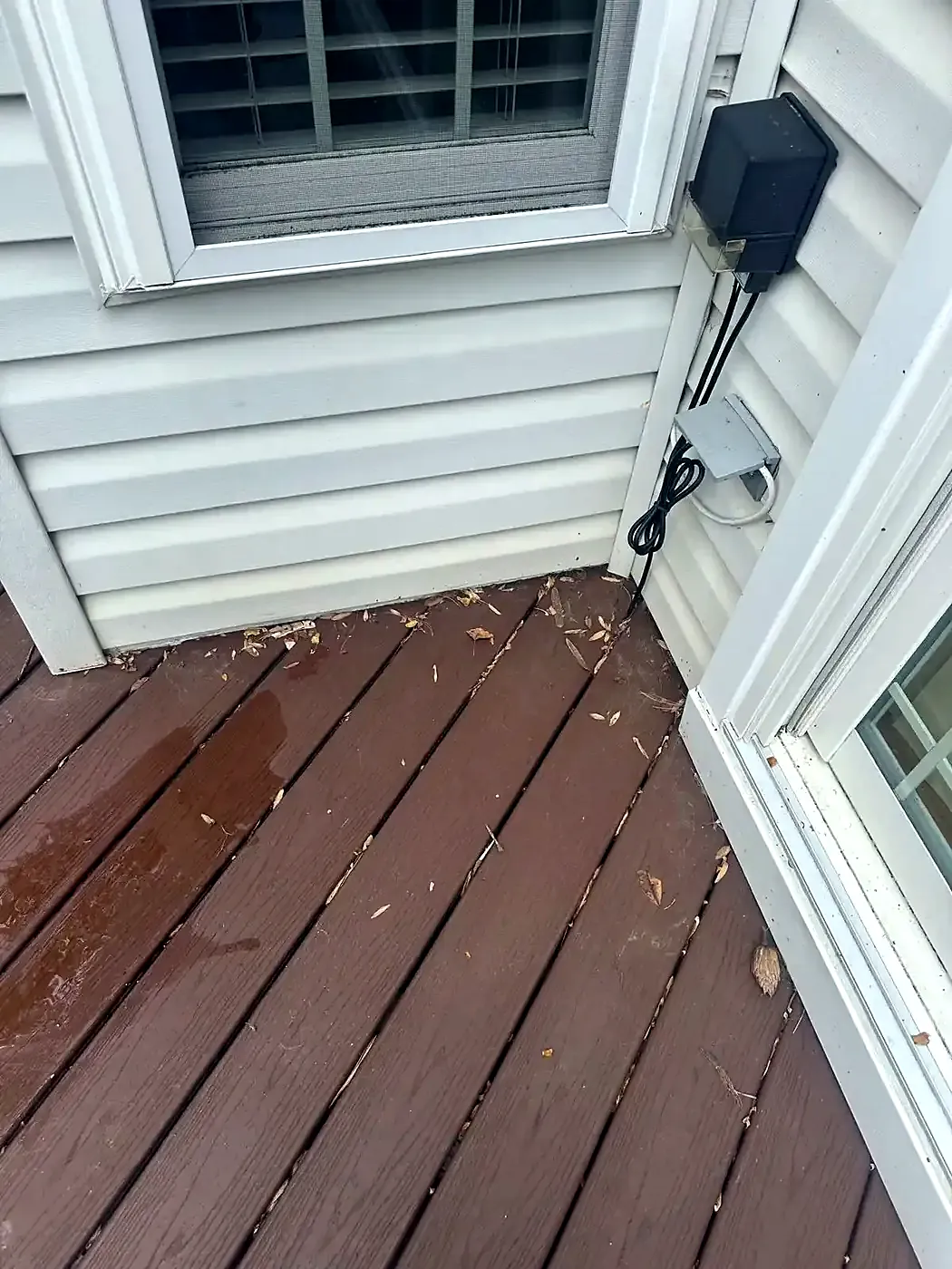 Worn brown deck boards outside a white door with leaf debris on the surface and siding above