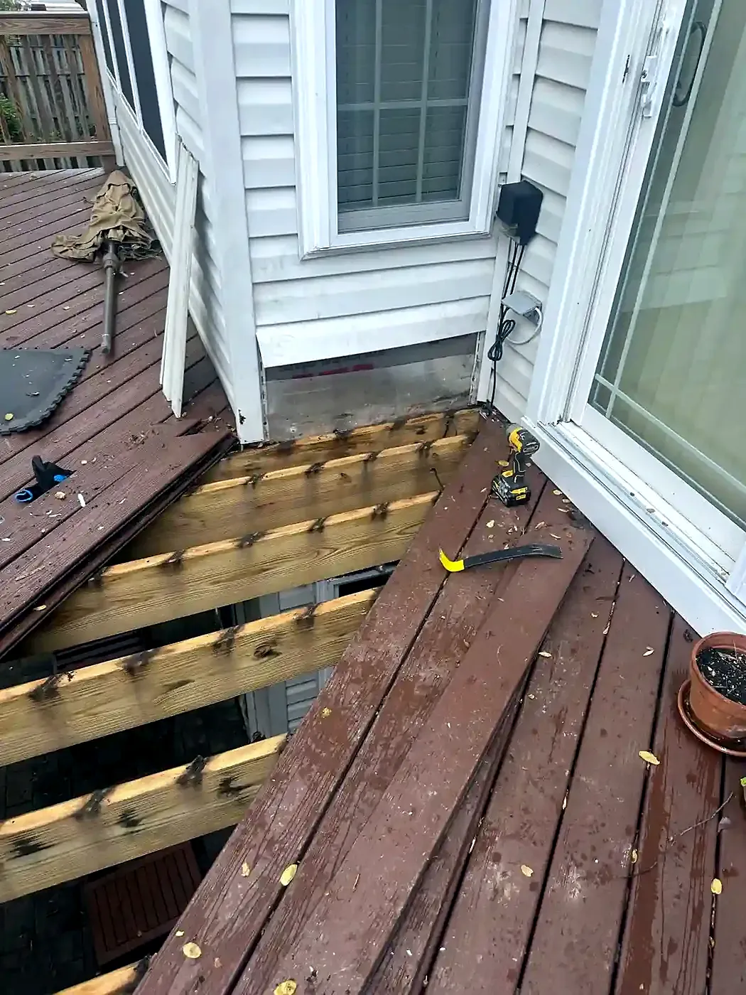 Deck boards pulled up beside a sliding door, exposing wood joists, with tools on the remaining planks