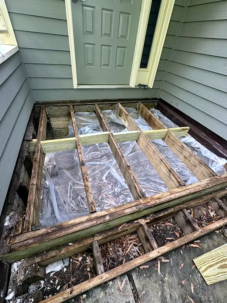 Weathered deck joists exposed under a door after the boards were removed, debris and plastic below