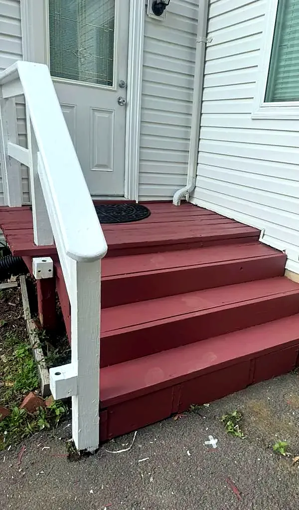 Red painted wood steps with a white handrail at a side door of a house with white siding