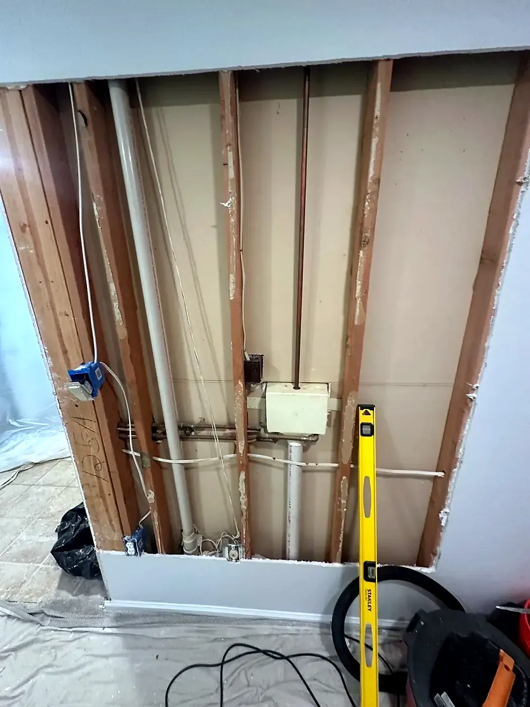 Interior wall stripped to bare studs showing pipes and electrical boxes, level leaning on the wall