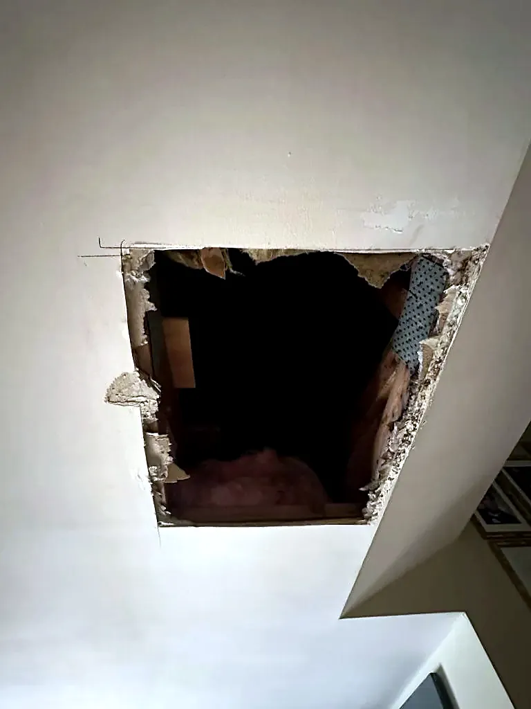 Ragged hole in a textured ceiling with framing and insulation visible above a stair wall