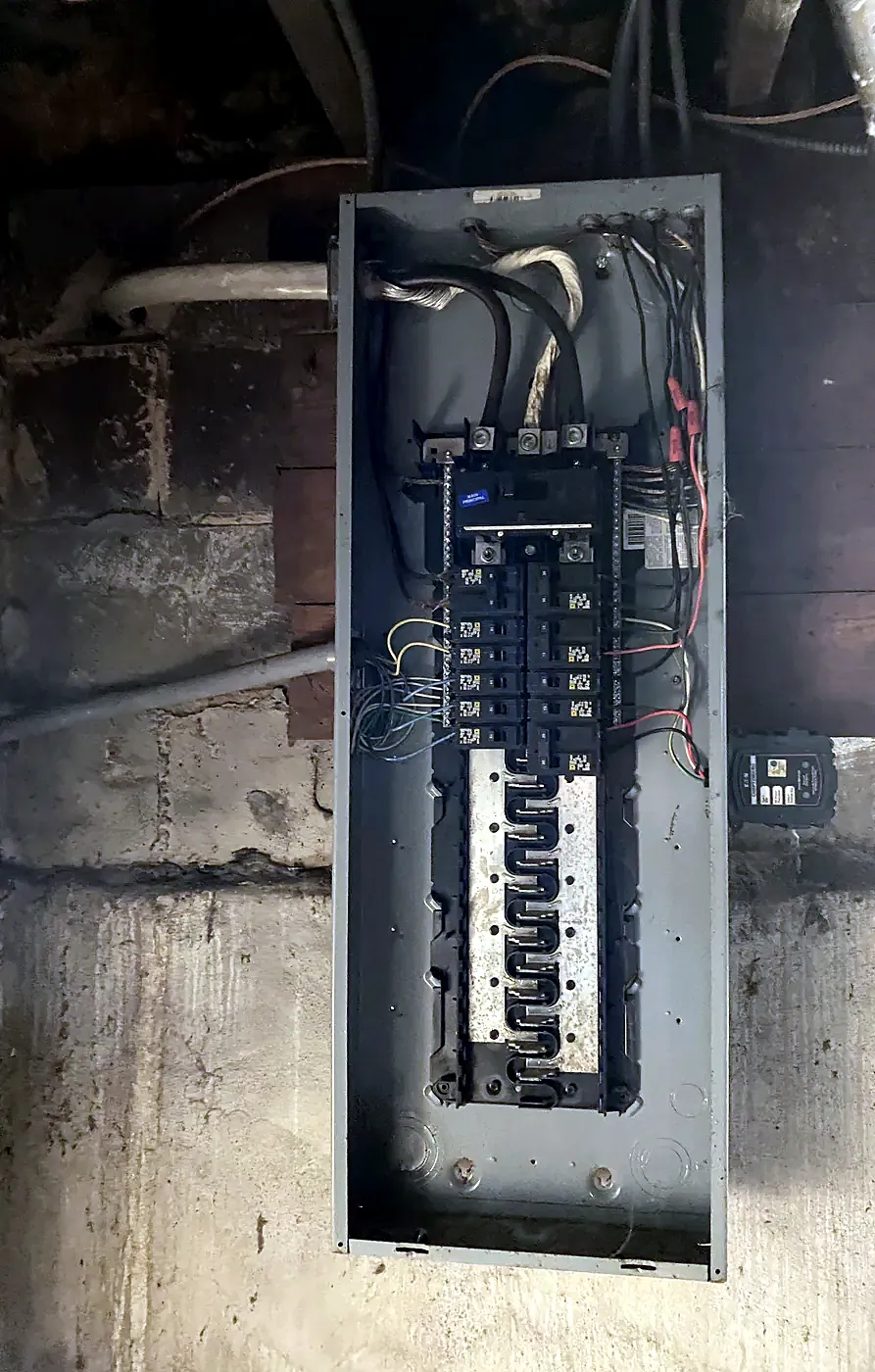 Old open electrical panel mounted on a dark, cobwebbed basement block wall