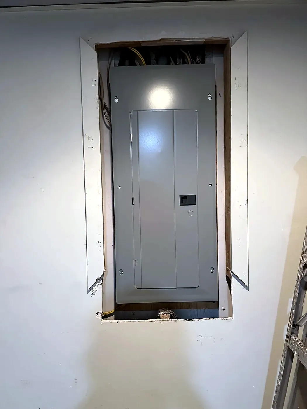 Closed gray electrical panel recessed into a white wall with rough drywall edges around the opening