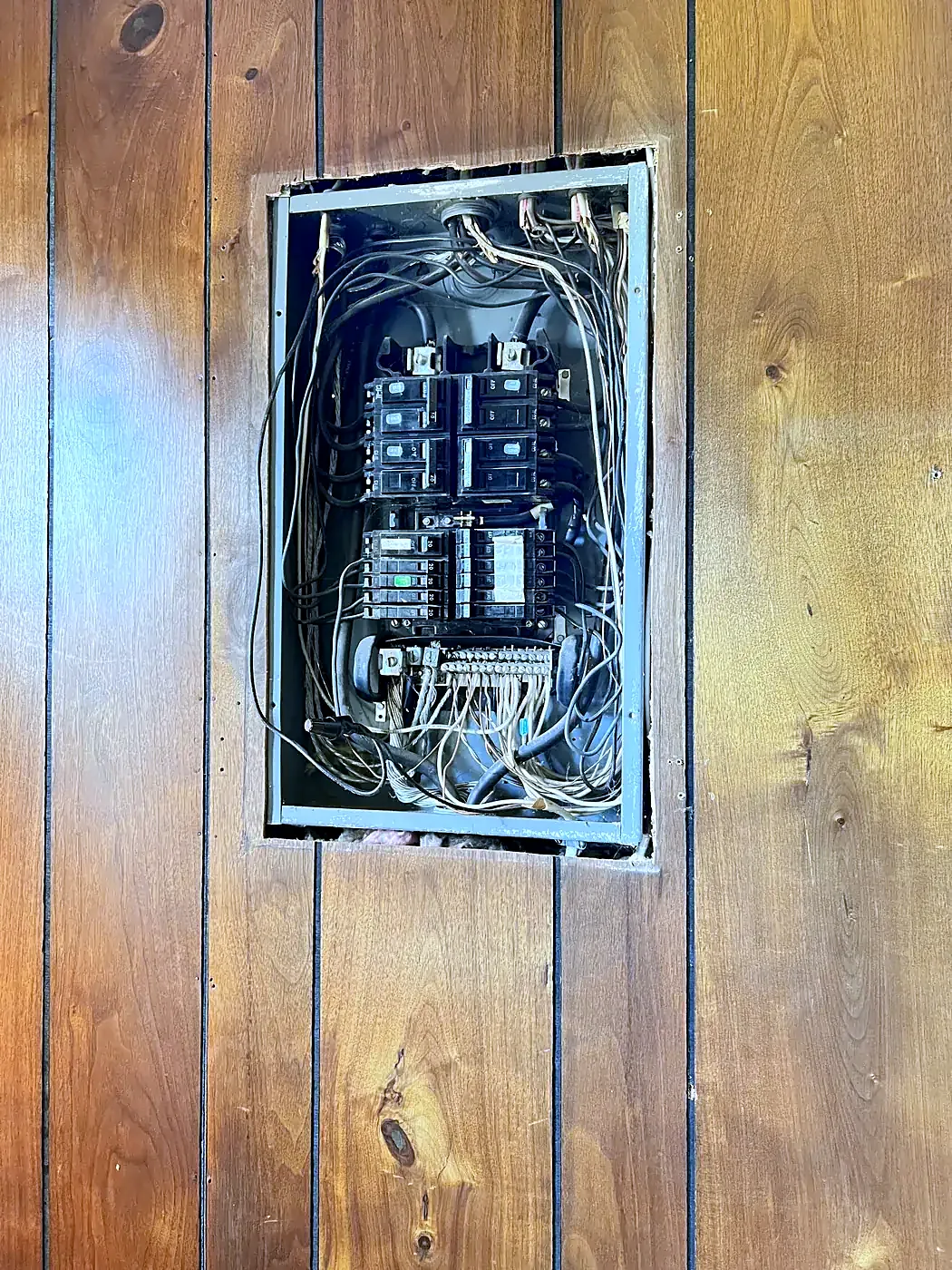 Open older electrical panel with exposed wiring set into a wall of knotty pine wood paneling