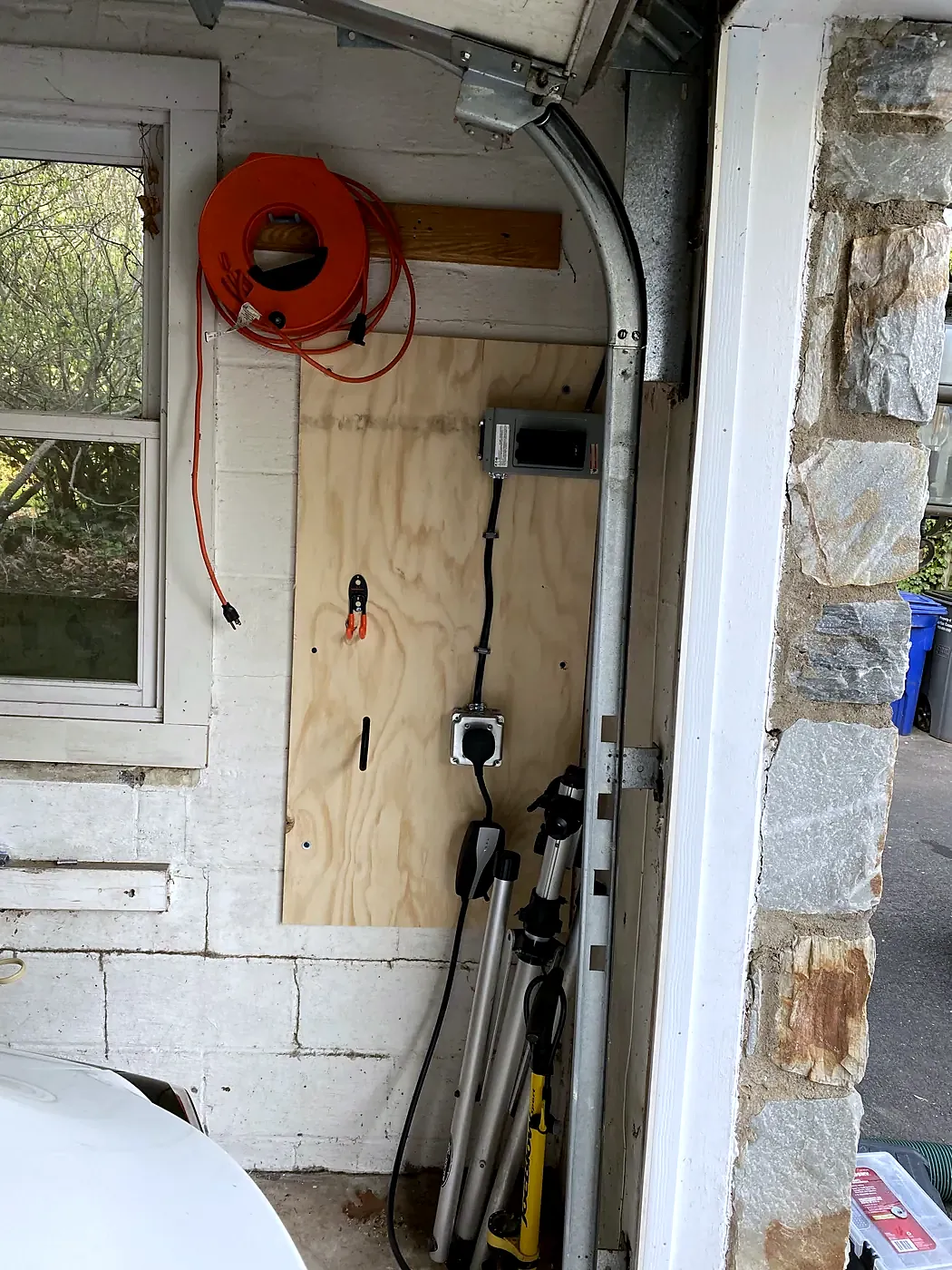 EV charger on a plywood-backed garage wall beside a window, stone pillar at the door opening