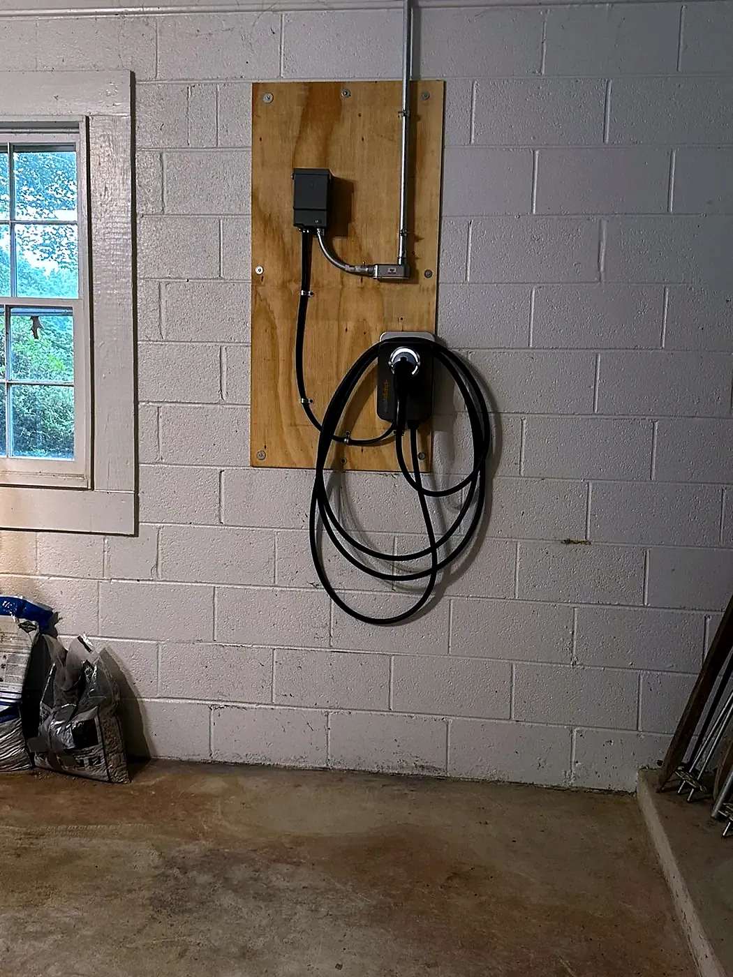 EV charger and conduit on a plywood board fastened to a cinderblock garage wall near a window