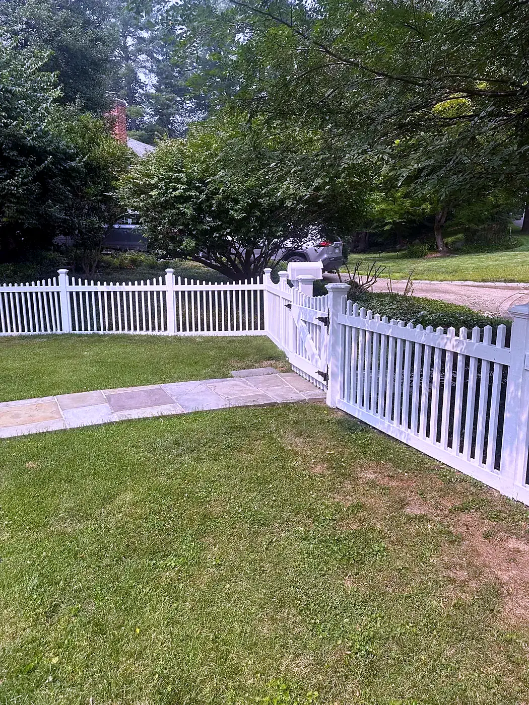White picket fence with a gate along a green lawn and flagstone walk, trees in the background