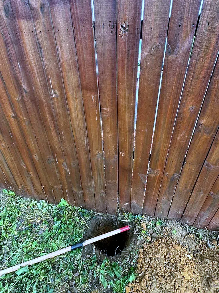 Freshly dug post hole with a digging bar beside a stained wood privacy fence