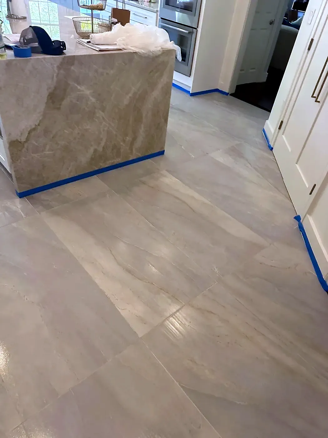 Large-format beige porcelain floor tiles laid in a kitchen with blue tape along the base of a stone island