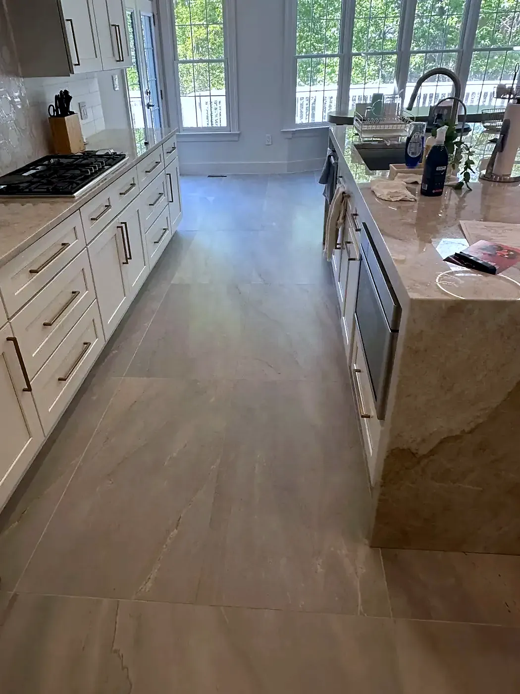 Finished beige porcelain tile floor in a white kitchen with a stone island, gas cooktop and window over the sink