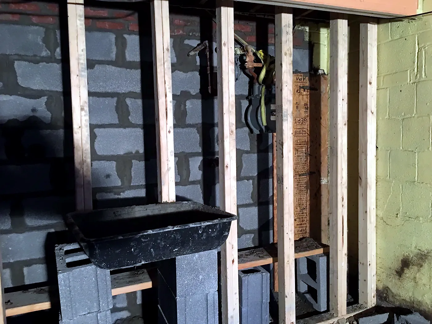 Gray concrete block basement wall behind exposed wood studs, black mixing tub on a stand in front