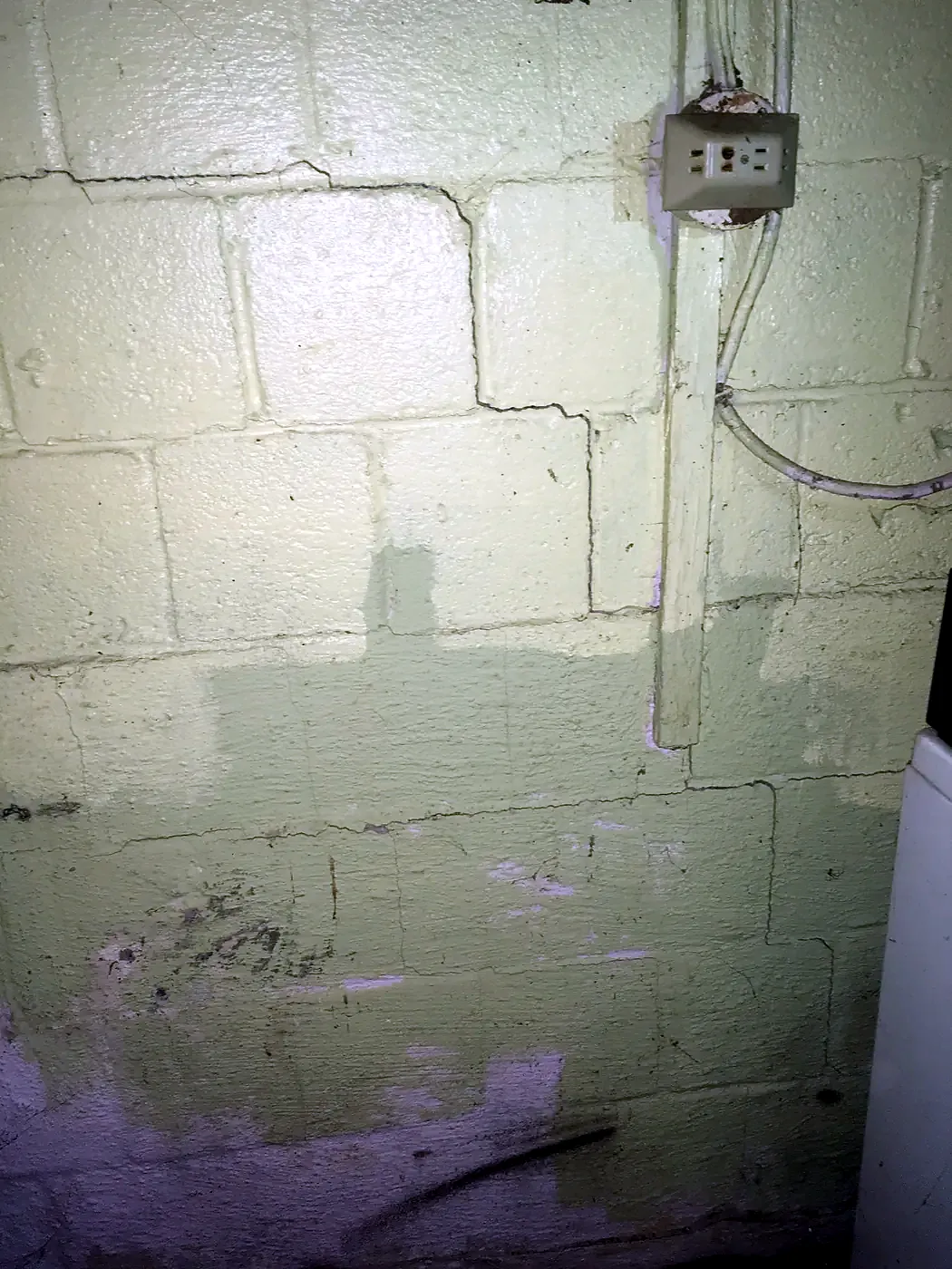 Yellow-painted concrete block basement wall with stair-step cracks along the mortar joints and an outlet box
