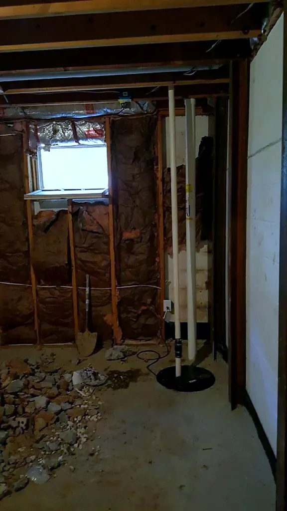 Unfinished basement with exposed studs, a small window and piles of rubble on the concrete floor