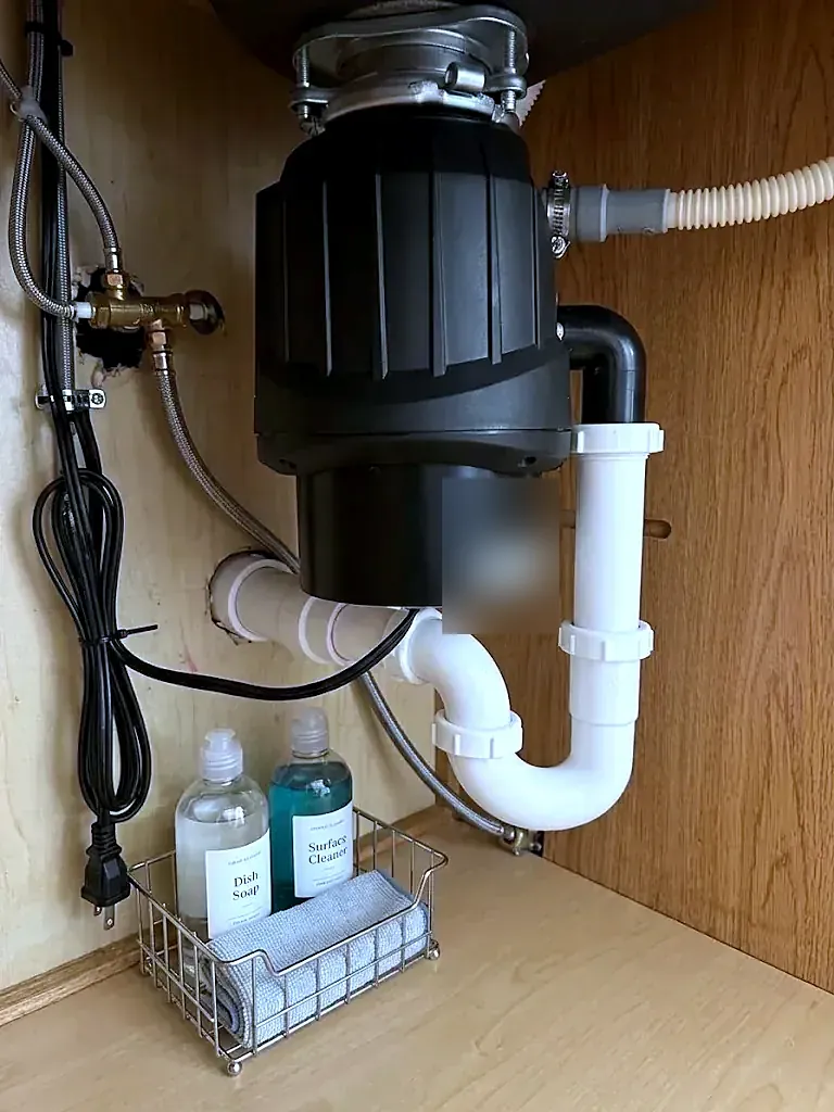 Black garbage disposal under a sink with white PVC drain pipe, dishwasher hose and soap bottles in a caddy