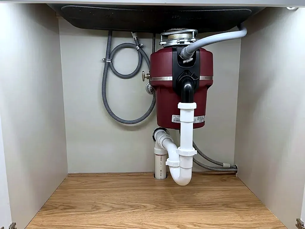 Dark red garbage disposal under a sink with white PVC trap and gray dishwasher hose in a clean cabinet