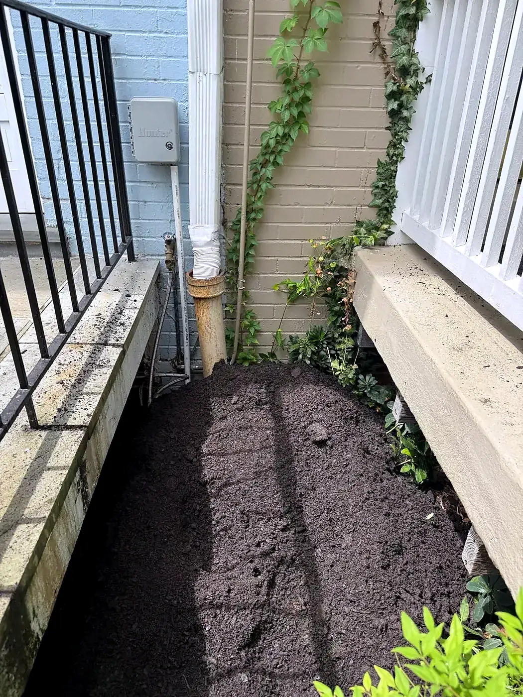 Narrow planter bed with fresh dark soil beside a house wall, white downspout and metal porch railing above