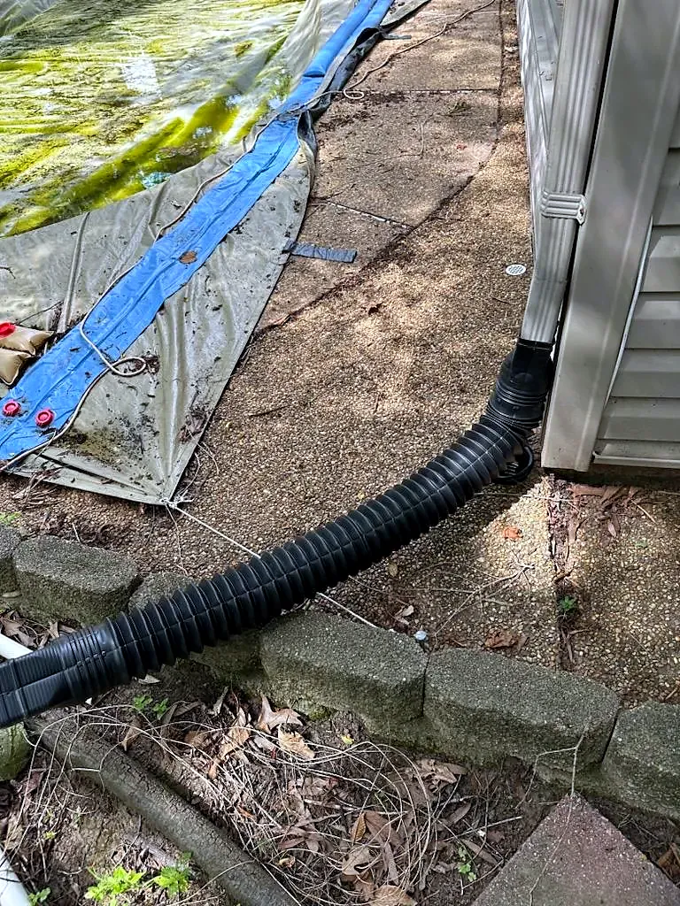 Black corrugated drain pipe running from a downspout along a block retaining wall, blue tarp on the ground