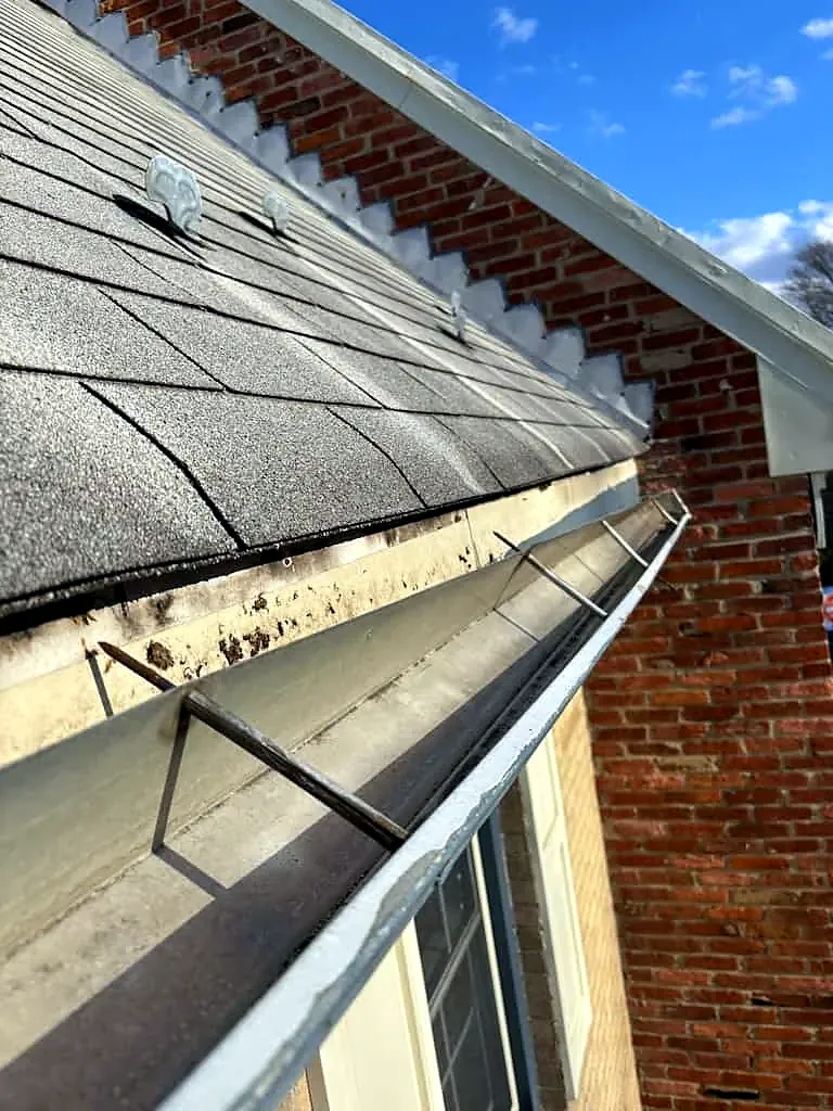 View along a gutter with metal hangers beneath the shingle roof edge on a brick house, blue sky above
