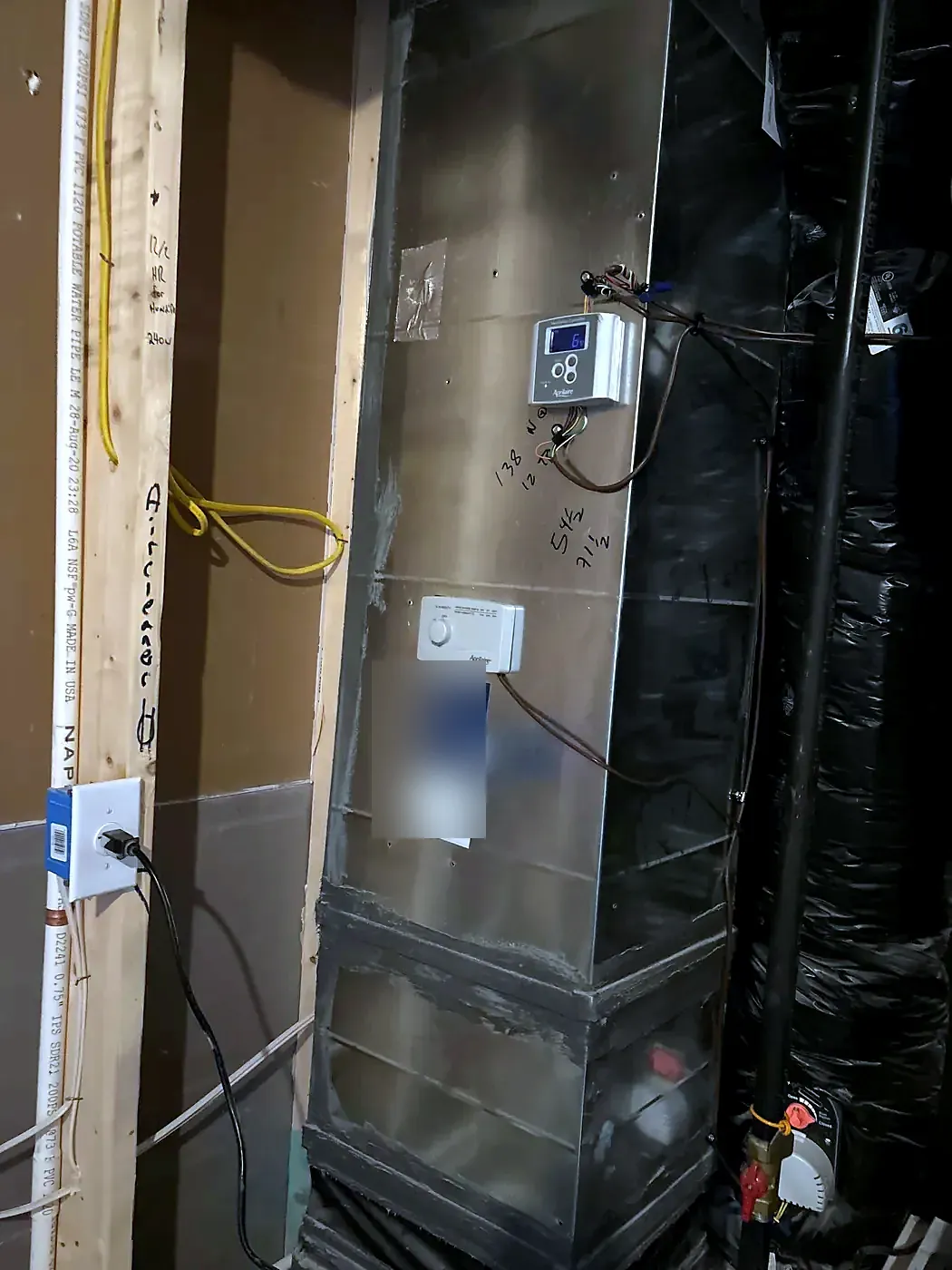Tall metal air handler with insulated black ductwork and a wall-mounted control beside it