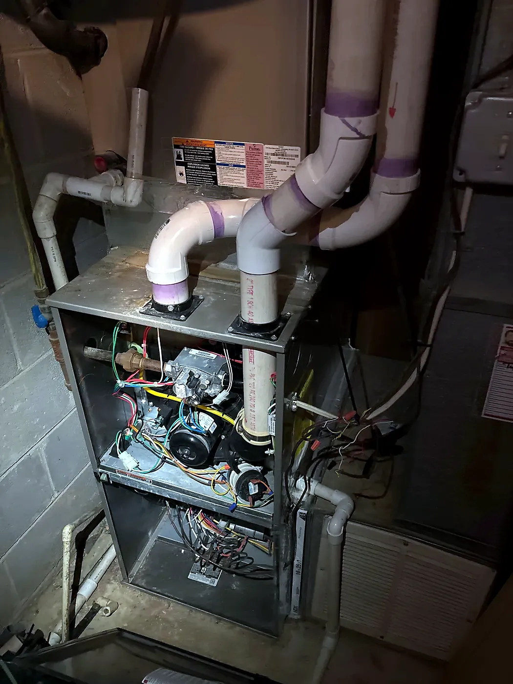 Furnace with panels off showing blower and wiring, PVC vent pipes with purple primer, block basement wall