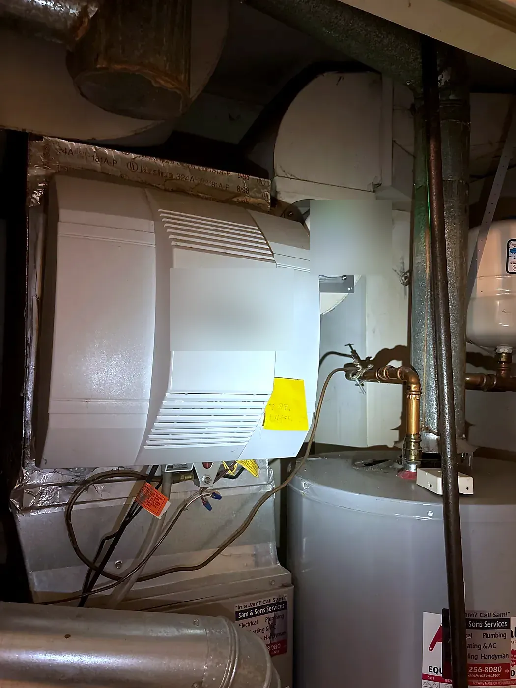 Beige whole-house humidifier unit installed on sheet metal ductwork next to a water heater, copper lines below