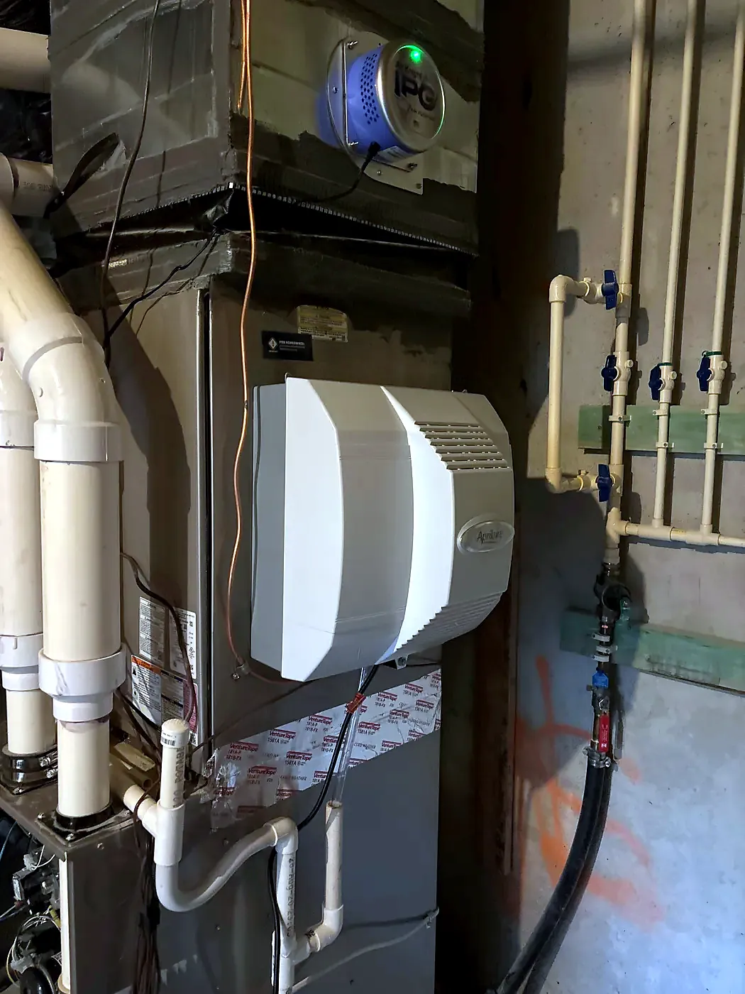 White humidifier on the side of a gray furnace with white PVC pipes and copper lines along a block wall