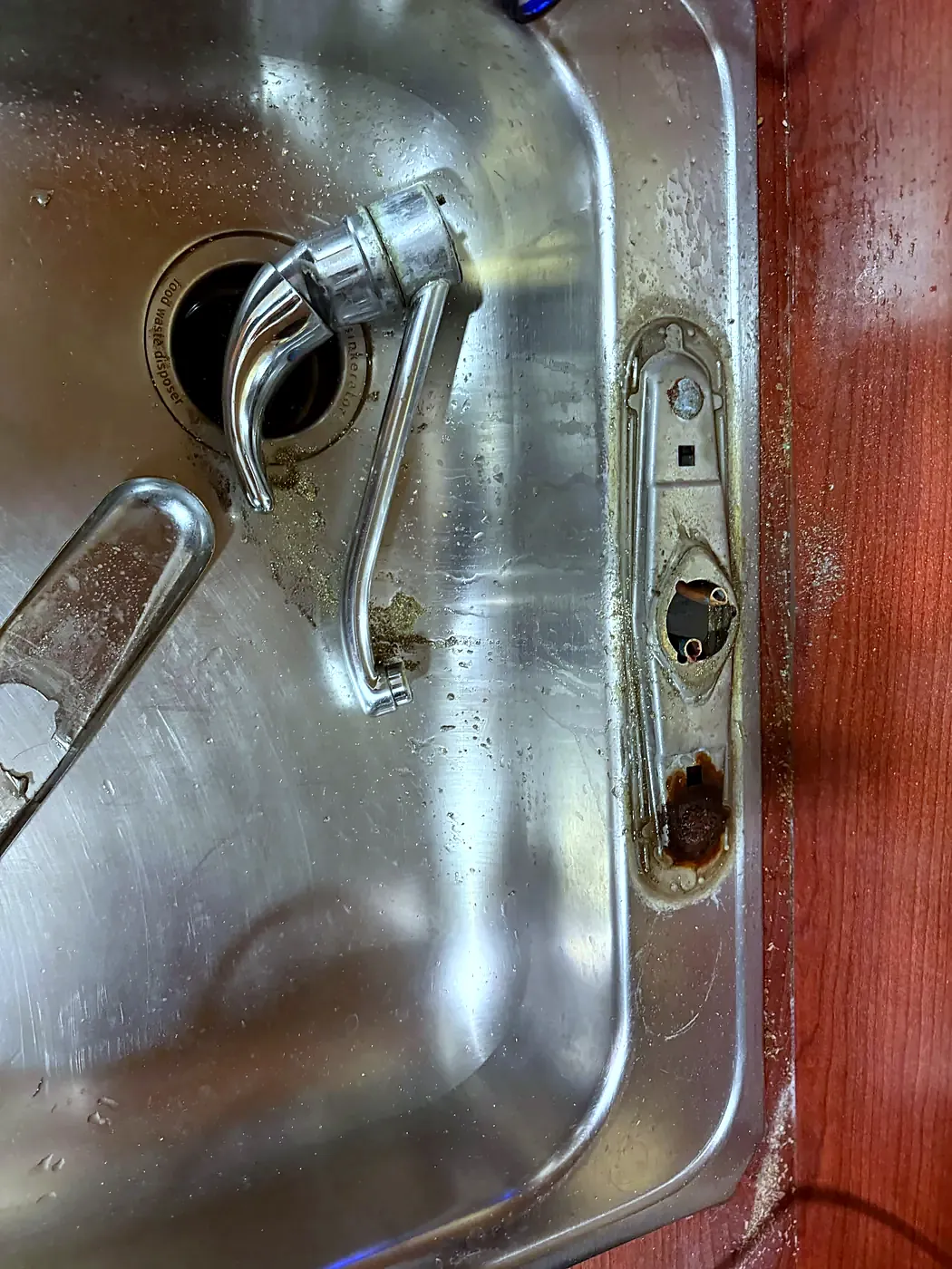 Old chrome kitchen faucet lifted off a corroded stainless sink deck with a red countertop