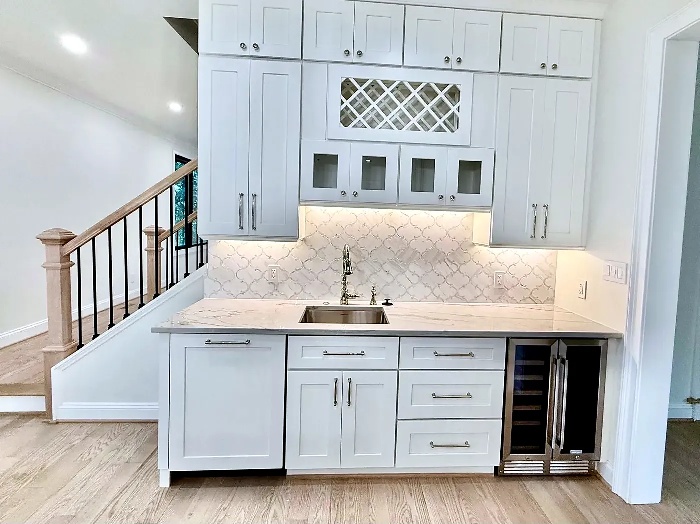 White bar cabinets with glass doors, a wine rack, small sink and wine fridge beside a staircase